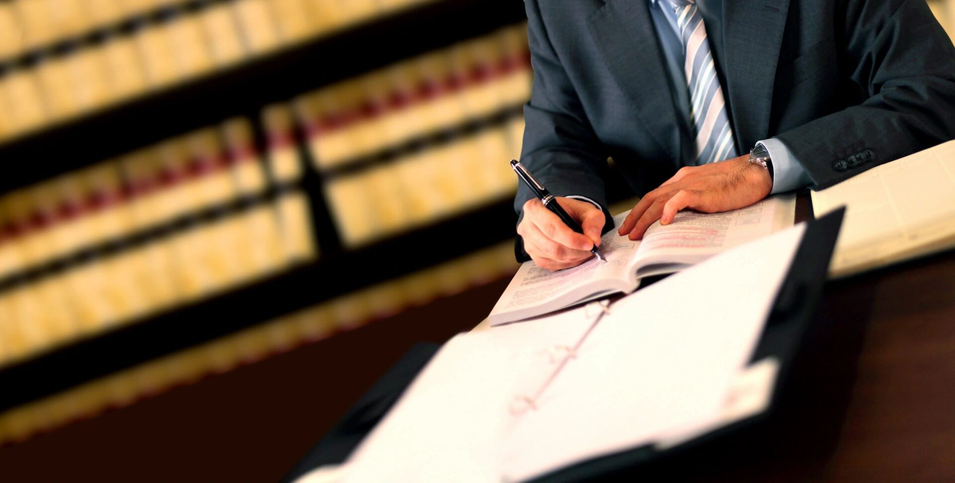 A person writing on paper at table in front of bookcase.